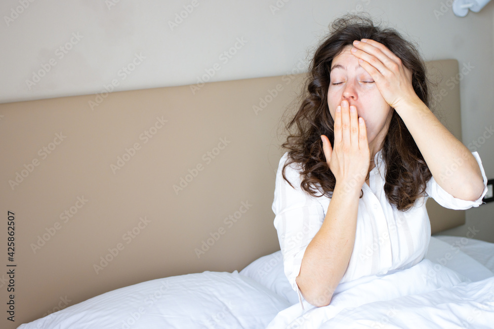Sleepy exhausted young brunette woman waking up in white bed at home, yawning covering her mouth, feeling headache or migraine, hangover. Suffering from insomnia, lack of sleep in morning. Copy space