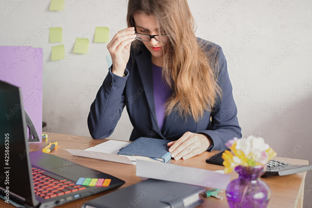 Fototapeta premium A beautiful young business woman is sitting at a desk, smiling and looking at a notebook. A natural working moment for a businesswoman in jacket. An open laptop, a diary, stationery. Working moment