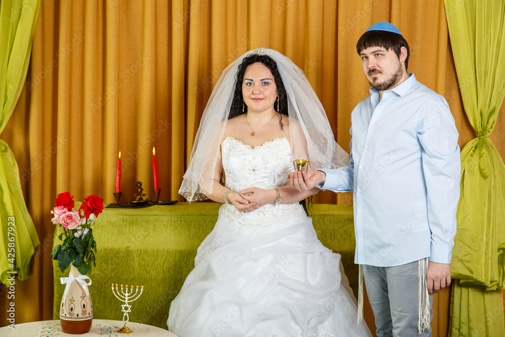 During the chuppah ceremony in the synagogue, the groom holds a glass of wine in his hand, the bride stands nearby.
