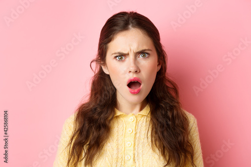 Shocked and hurt young woman frowning, staring offended and disappointed, gasping, looking judgemental, standing over pink background photo