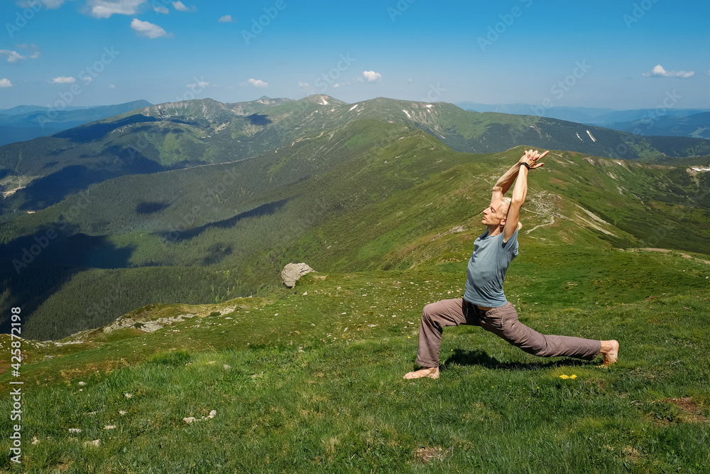 Strong man practicing yoga exercises under sky on mountain top