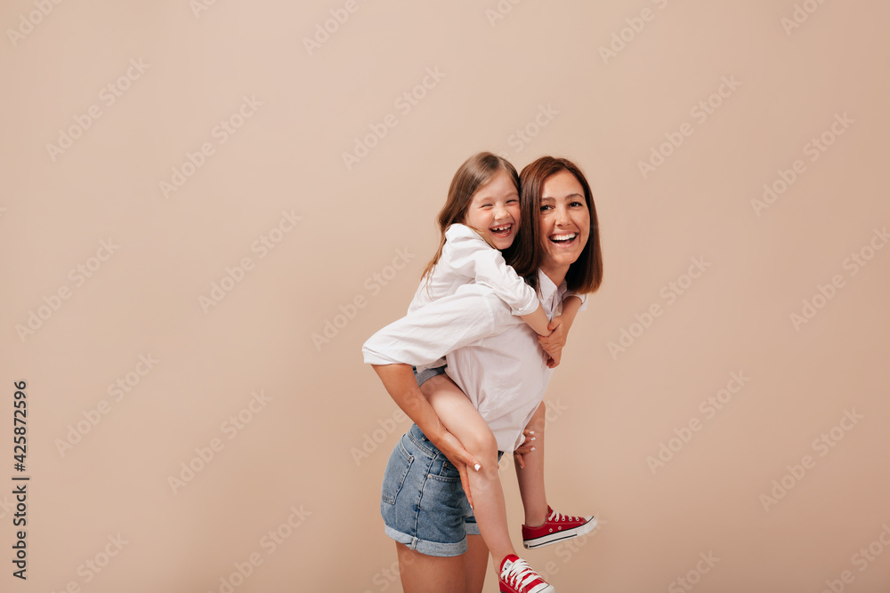 Smiling happy woman in white shirt and shorts holding her daughter on her back and smiling at camera over beige backdrop Smiling happy woman in white shirt and shorts holding her daughter on her back and smiling at camera over beige backdrop