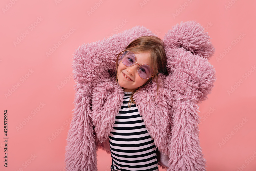 Little charming girl wearing fur trendy coat and round glasses is throwing hands up and smiling at camera over pink backdrop Little charming girl wearing fur trendy coat and round glasses is throwing hands up and smiling at camera over pink backdrop