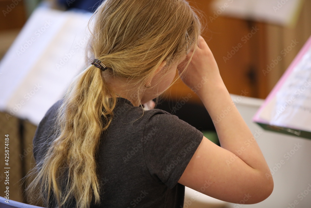 Fototapeta premium Portrait of a little girl with long hair gathered in a ponytail sits with a raised hand at her head and touches her forehead at a music lesson in class with notes.Tired child.Back view.Close-up