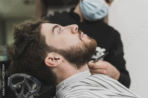A young man fixes his beard at the barbershop. The barber wears a protective mask against the spread of the coronavirus. The so-called new normal during the 2020-21 pandemic