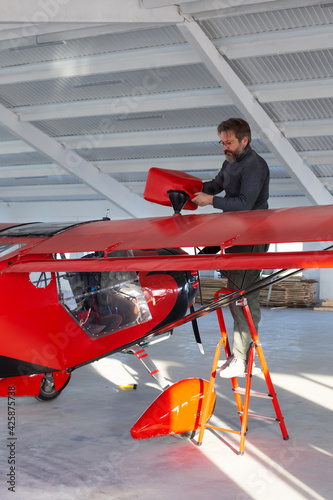 Single engine aircraft, ultra light plane before flight. Pilot is refueling small motor airplane by red jerrican at hangar in private airport. Private aerodrome. Red air vehicle and ladder.