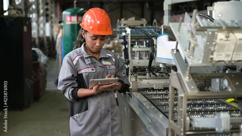 A young black manufacturing worker controls an assembly line in a factory, making notes on a tablet. Young woman in hard hat watches the work process