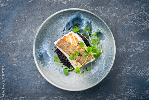 Modern style traditional fried skrei cod fish filet with portulaca lettuce, and black rice served as top view on ceramic design plate with copy space photo