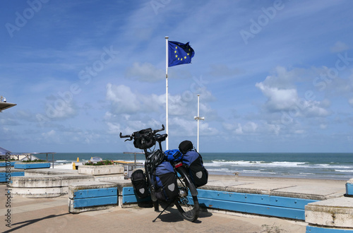 A packed touring bike on the French coast in front of a waving European flag.