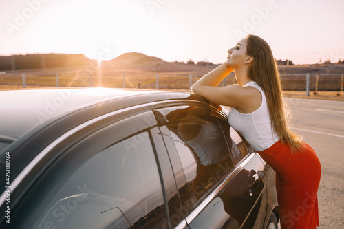 Beautiful young girl near a car in the evening in the sunset sun in an empty parking lot © Daria Lukoiko