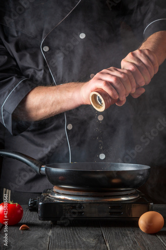 The chef adds pepper while cooking eggs in pan. Work environment on vintage kitchen table. Vertical image