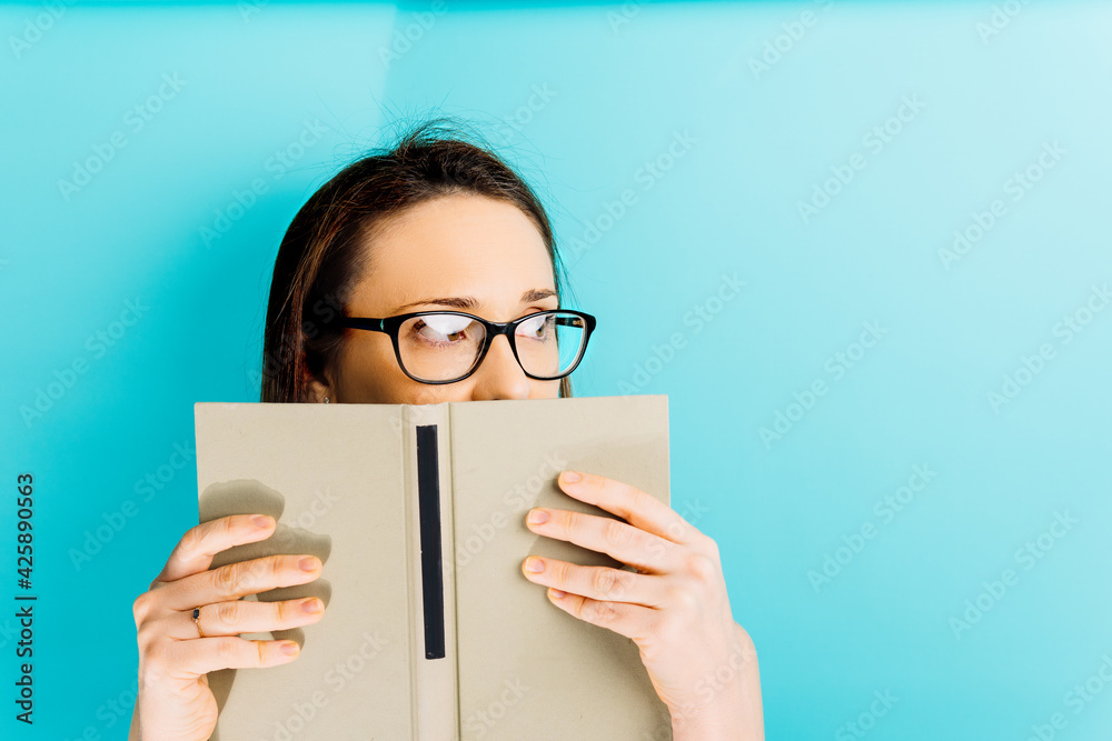 Beautiful young woman covering her face with a book while looking aside with space for copy. blue background. reading concept Beautiful young woman covering her face with a book while looking aside with space for copy. blue background. reading concept