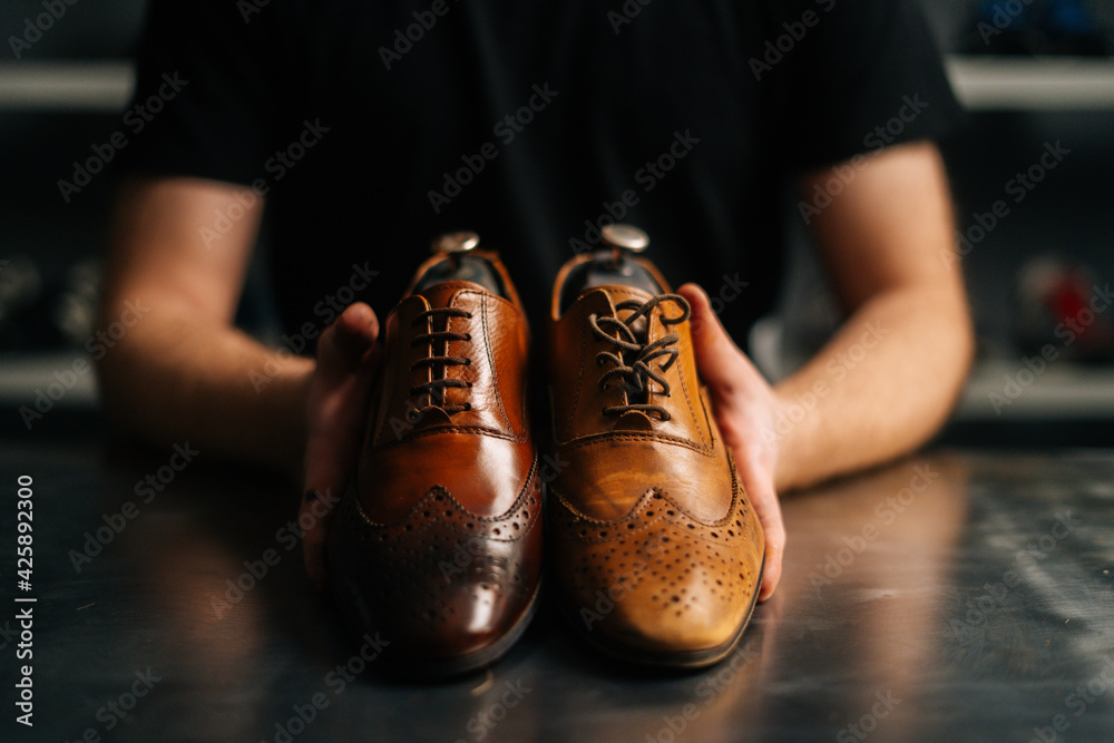 Close-up hands of male shoemaker holds old light brown leather shoe and  repaired shiny shoes after restoration working. Concept of cobbler artisan  repairing and restoration work in shoe repair shop. Stock Photo