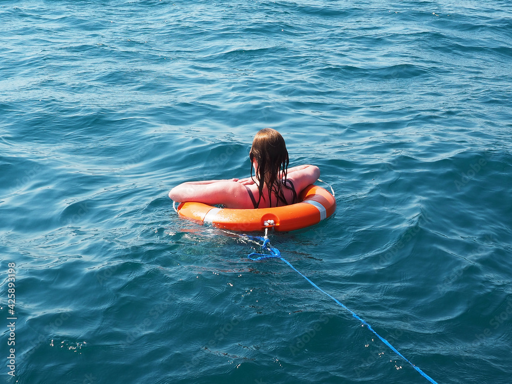 Girl in an orange lifebuoy with a rope swims in the open blue sea on a sunny day