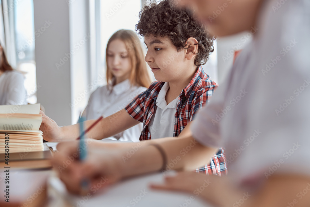 Young and curly schoolboy sitting on desk and writing. Elementary school kids sitting on desks and reading books in classroom