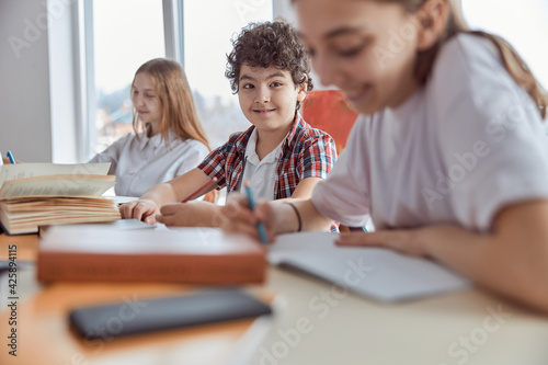 Young and curly schoolboy sitting on desk and smiling. Elementary school kids sitting on desks and reading books in classroom