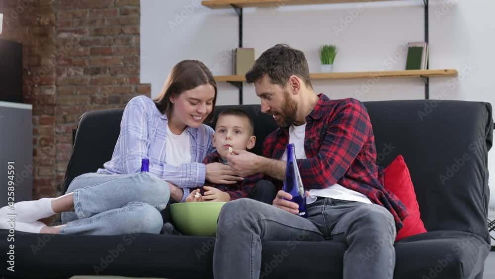 Happy young parents with cute baby sit on sofa in living room at home and watch funny movie, eat popcorn and spend weekend together Happy young parents with cute baby sit on sofa in living room at home and watch funny movie, eat popcorn and spend weekend together