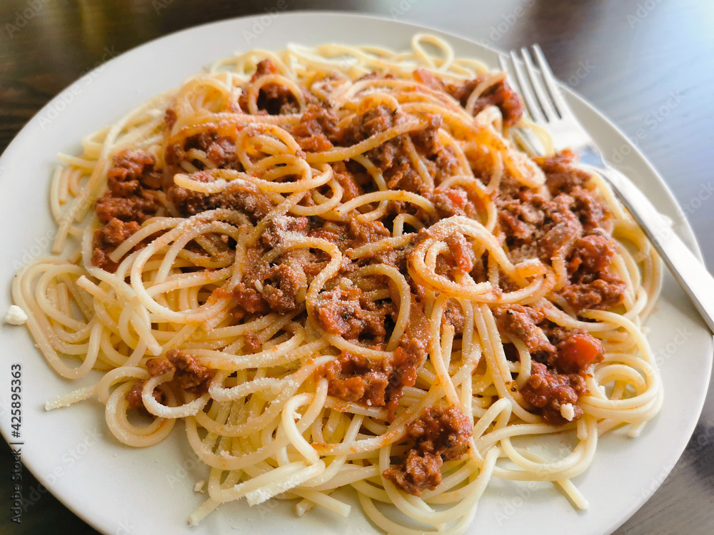 Spaghetti Bolognese with minced beef, onion, chopped tomato, garlic, olive oil, stock cube, tomato puree and Italian herb. Traditional Italian food on a white plate with a fork on the right. Spaghetti Bolognese with minced beef, onion, chopped tomato, garlic, olive oil, stock cube, tomato puree and Italian herb. Traditional Italian food on a white plate with a fork on the right.