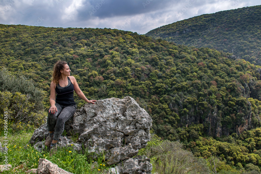 woman traveler sitting on rock and looking at amazing green hills and forest, hiking trip through Israel mountains, wanderlust travel concept, space for text, atmospheric epic moment