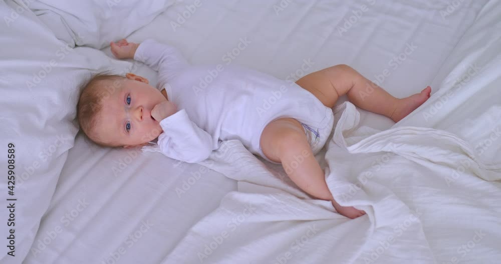 Newborn baby in a white bodysuit sucks fingers in his mouth, lies on a bed in a bright room. Cute baby with big blue eyes, childhood. Top view of the camera. 4k, ProRes Newborn baby in a white bodysuit sucks fingers in his mouth, lies on a bed in a bright room. Cute baby with big blue eyes, childhood. Top view of the camera. 4k, ProRes