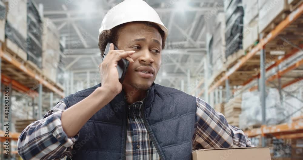 Close up portrait of young African American handsome guy engineer specialist talking on smartphone standing at work at construction site in helmet. Male worker calling on cellphone. Factory concept
