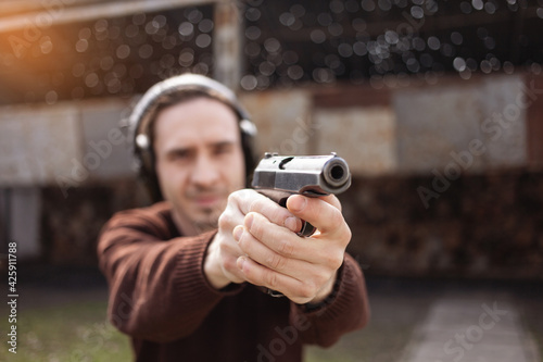 A man shoots a gun, aiming at the target. A man wearing protective headphones. A wall and a roof with bullet holes. outdoor shooting range