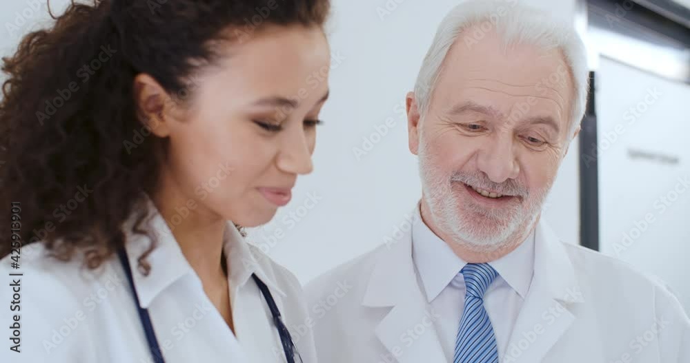 Portrait two medical workers aged caucasian male and young curly hair multiethnic female, doctors discussing medical case, talking about health report while standing by white wall in hospital Portrait two medical workers aged caucasian male and young curly hair multiethnic female, doctors discussing medical case, talking about health report while standing by white wall in hospital