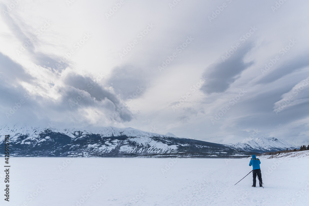 Man in a blue jacket standing on a frozen lake photographing, taking photos of the stunning, snowy, winter landscape with clouds shining above. 