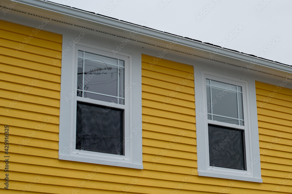 A bright yellow cape cod horizontal siding wood exterior wall with two glass double hung windows and white eve. The background is a grey cloudy sky. A bright yellow cape cod horizontal siding wood exterior wall with two glass double hung windows and white eve. The background is a grey cloudy sky.