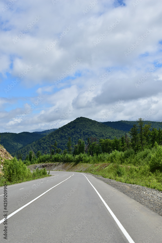 Fototapeta premium Asphalt road in the hills. Among the green mountains is a road. Around green hills, dense forest, blue sky. The Sikhote-Alin Mountains.
