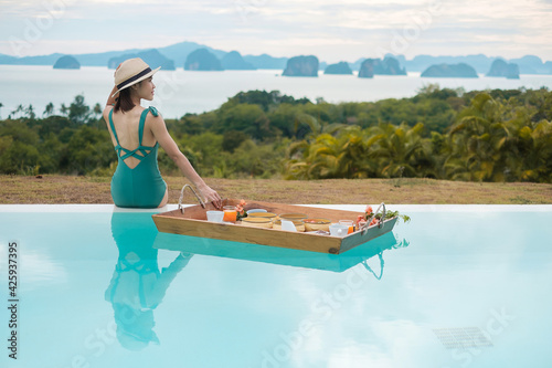 Happy young woman in green swimsuit having Floating Breakfast tray in luxury pool hotel, girl with hat enjoy in tropical resort. Relaxing, Exotic summer travel, holiday, vacation and tropical