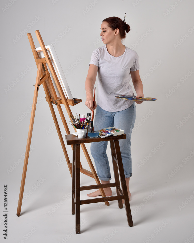 Full length portrait of a red haired artist girl wearing casual jeans and white shirt.  standing pose 
painting a canvas on an easel, against a studio background.