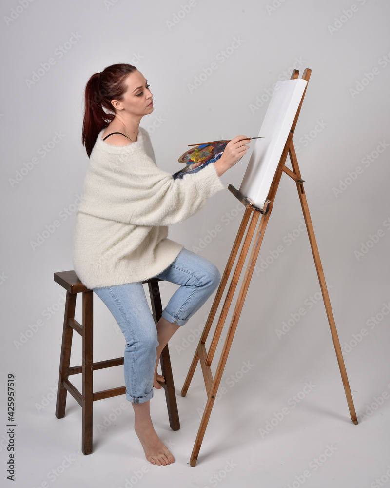 Fototapeta premium Full length portrait of a red haired artist girl wearing casual jeans and white shirt. Sitting pose on chair, painting a canvas on an easel against a studio background