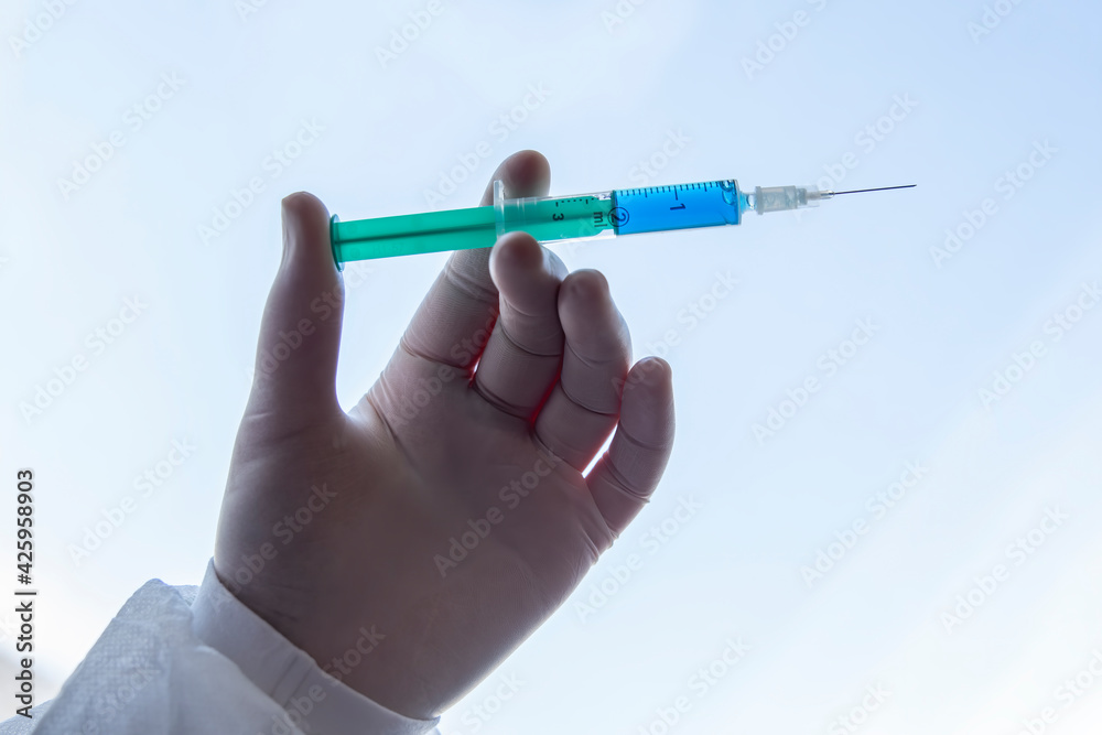 Doctor wearing surgical gloves holding a needle or syringe for injection with a blue liquid against white blue background. Doctor wearing surgical gloves holding a needle or syringe for injection with a blue liquid against white blue background.