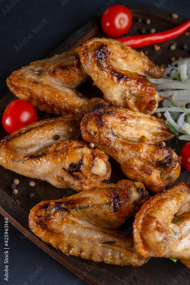 grilled on coals chicken wings on a wooden board with pickled onions, chili peppers and cherry tomatoes on a dark background. photos with a small depth of field, top view grilled on coals chicken wings on a wooden board with pickled onions, chili peppers and cherry tomatoes on a dark background. photos with a small depth of field, top view