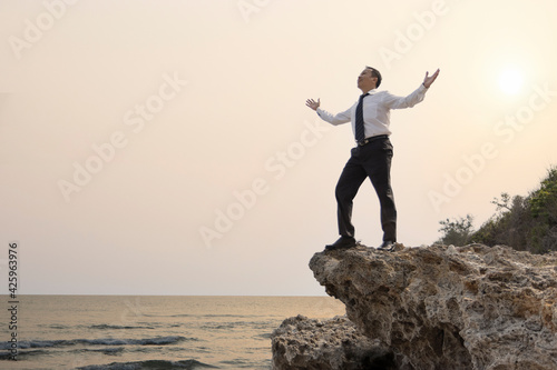 Asian businessman standing on the cliff with two arms rising showing of great pleasure or happy satisfaction of enjoyment when business target reached or hard job finished