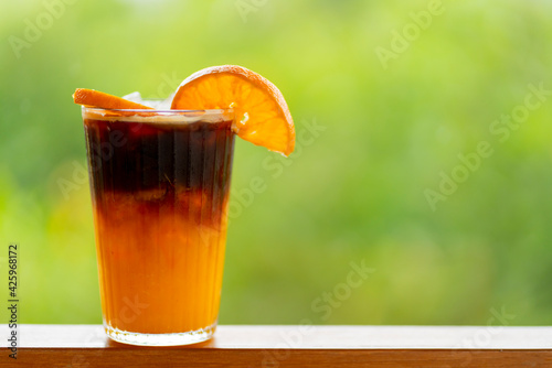 Americano coffee mixed with orange juice in a clear glass can be seen in layers. Behind it is a refreshing green bokeh.