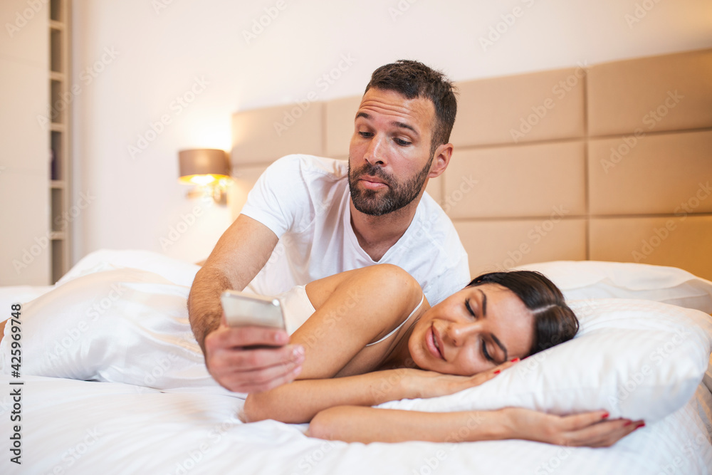 Jealous husband spying the phone of his partner while she is sleeping in a bed at home. Shocked jealous husband spying the phone of his wife while she sleeping in bed at home Jealous husband spying the phone of his partner while she is sleeping in a bed at home. Shocked jealous husband spying the phone of his wife while she sleeping in bed at home