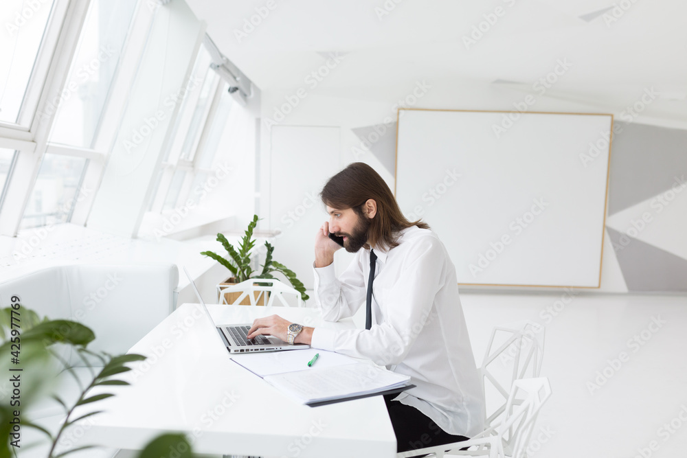 a business man in a white shirt and tie sits working with a laptop in a modern expensive office by the window
