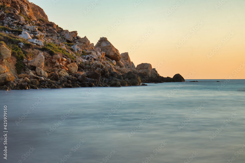 Orange sky over rocks and blue water at sunset at Golden Bay, Malta.