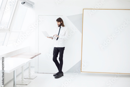 a man in a white shirt and tie holds a laptop in his hands leaning against a white wall. shot on a white background wall near the window  © Bilous