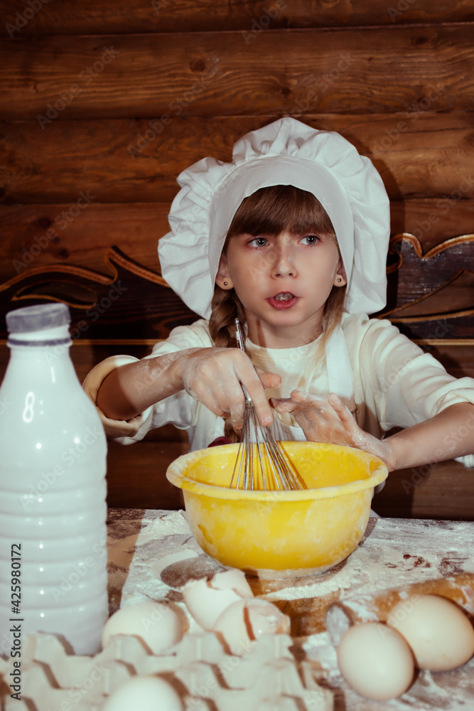 A little funny girl dressed as a cook kneads the dough. Homemade baking. Wooden background. Cooking. A little funny girl dressed as a cook kneads the dough. Homemade baking. Wooden background. Cooking.