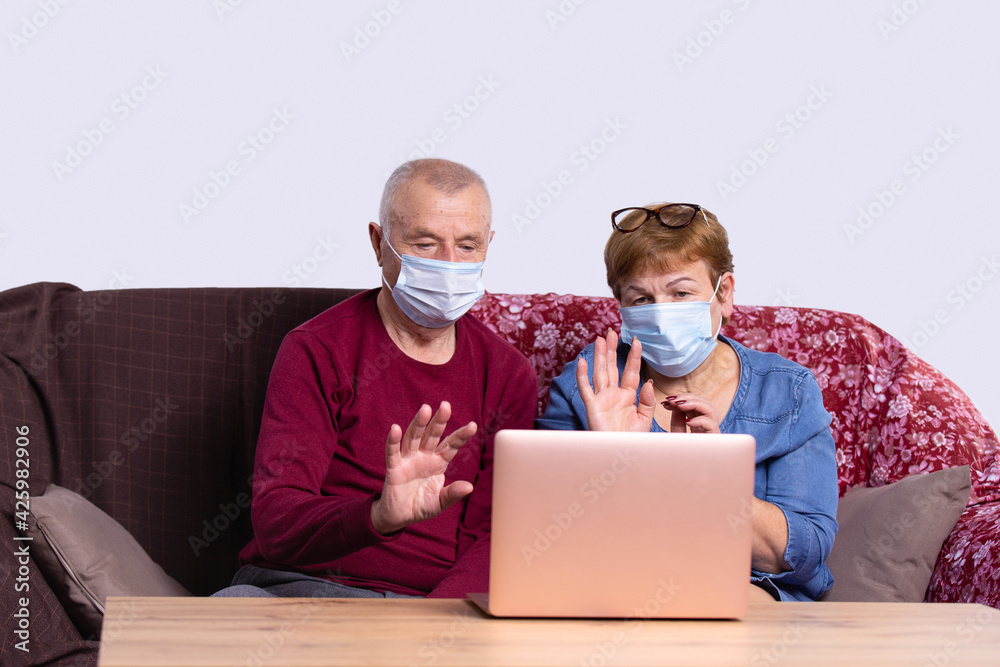 Couple old aged senior people at home with seasonal winter cold illness disease looking at laptop, gesturing hello, sit down on the sofa. Elderly couple in medical masks during the pandemic Couple old aged senior people at home with seasonal winter cold illness disease looking at laptop, gesturing hello, sit down on the sofa. Elderly couple in medical masks during the pandemic