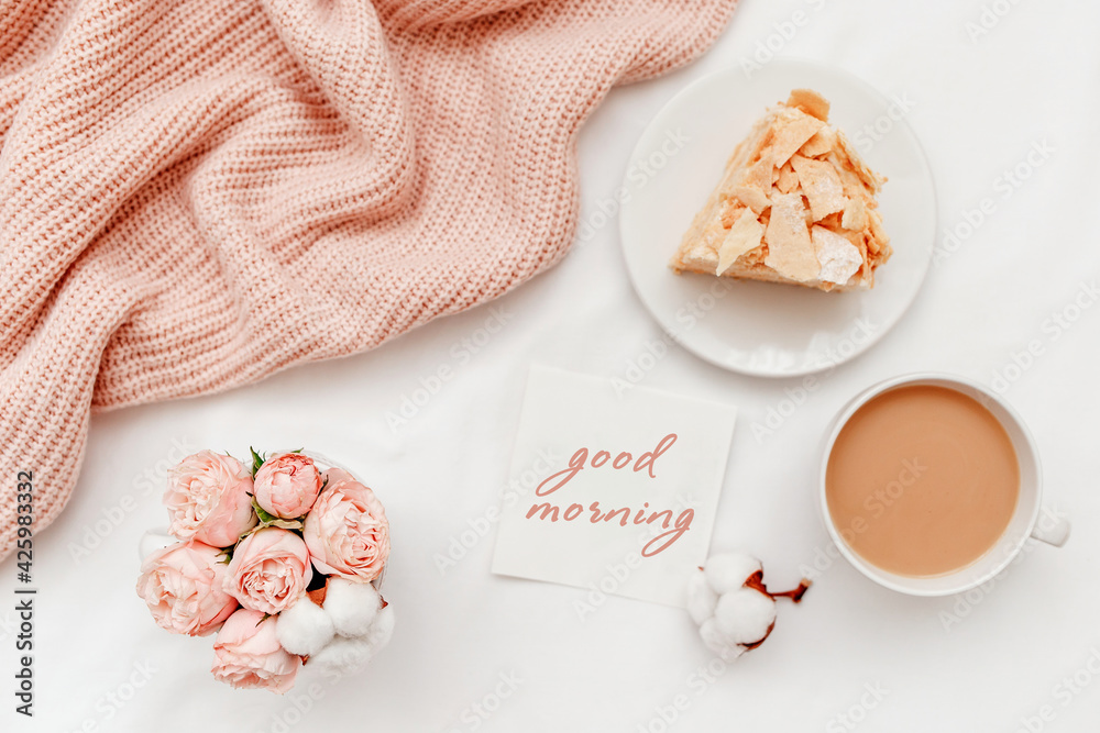 Cup of coffee with milk, piece of cake, rose flowers, pink knitted plaid or blanket, card with text GOOD MORNING. Breakfast in bed. Stylish home interior decor. Flat lay, top view. Cup of coffee with milk, piece of cake, rose flowers, pink knitted plaid or blanket, card with text GOOD MORNING. Breakfast in bed. Stylish home interior decor. Flat lay, top view.
