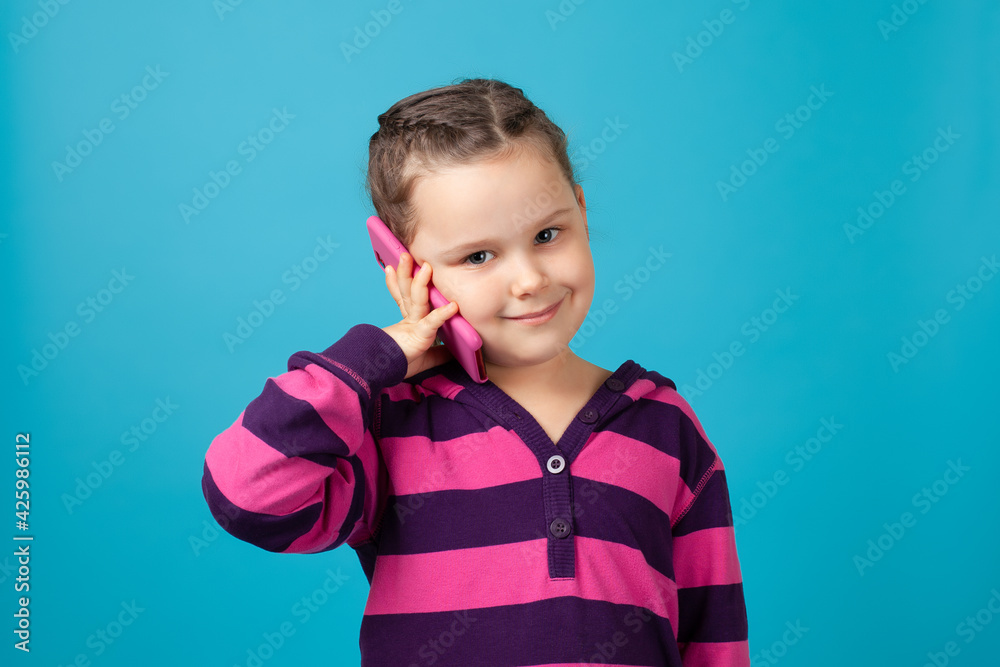 close-up portrait of a smiling beautiful child with pigtails holding a phone near her ear and making a phone call, isolated on a blue background.