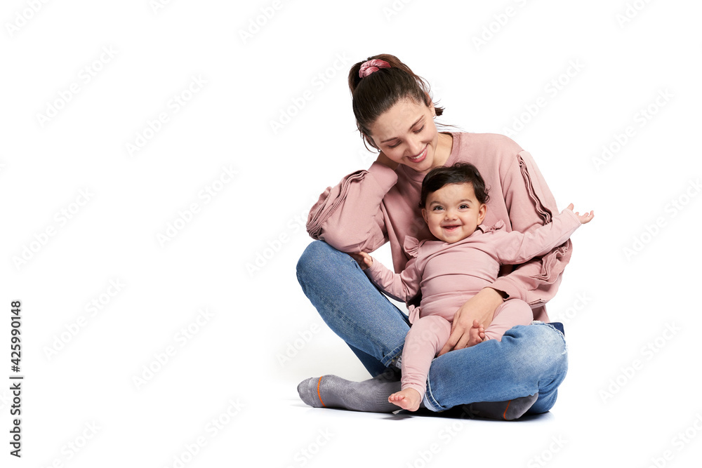 Portrait of cute mother with daughter posing isolated on white. Young woman holding sweet adorable baby girl on leg, looking at child and touching neck while sitting on floor in lotus pose.