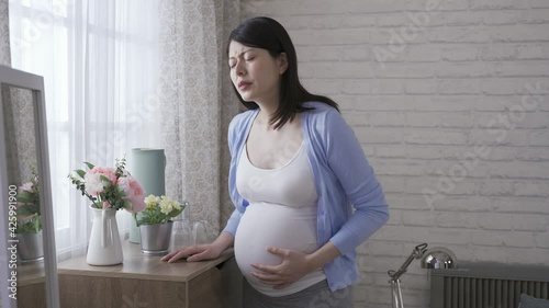 chinese pregnant mom approaching to window is touching her belly while suffering unexpected contraction pain in the bedroom.