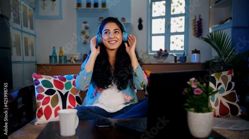 Cute girl in her early twenties listening to her favorite music sitting on the sofa. Cheerful Indian female taking a break from work and relaxing while hearing peppy songs using her blue headphones