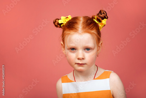 A little girl with red hair stands on a pink background and smiles.