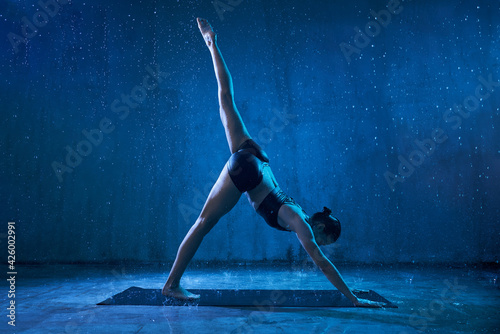 Sporty attractive woman wearing black sportswear standing in yoga asana with raised leg under rain. Strong flexible girl standing in downward facing dog, adho mukha svanasana pose. photo